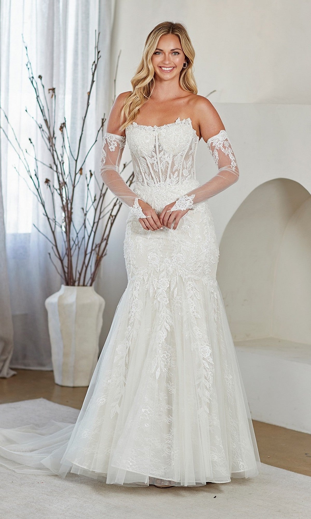 A woman wearing the white Amelia Couture 2155 long prom dress stands indoors, smiling at the camera. The strapless, fitted gown features lace embroidery, a corset bodice, and sheer long gloves.
